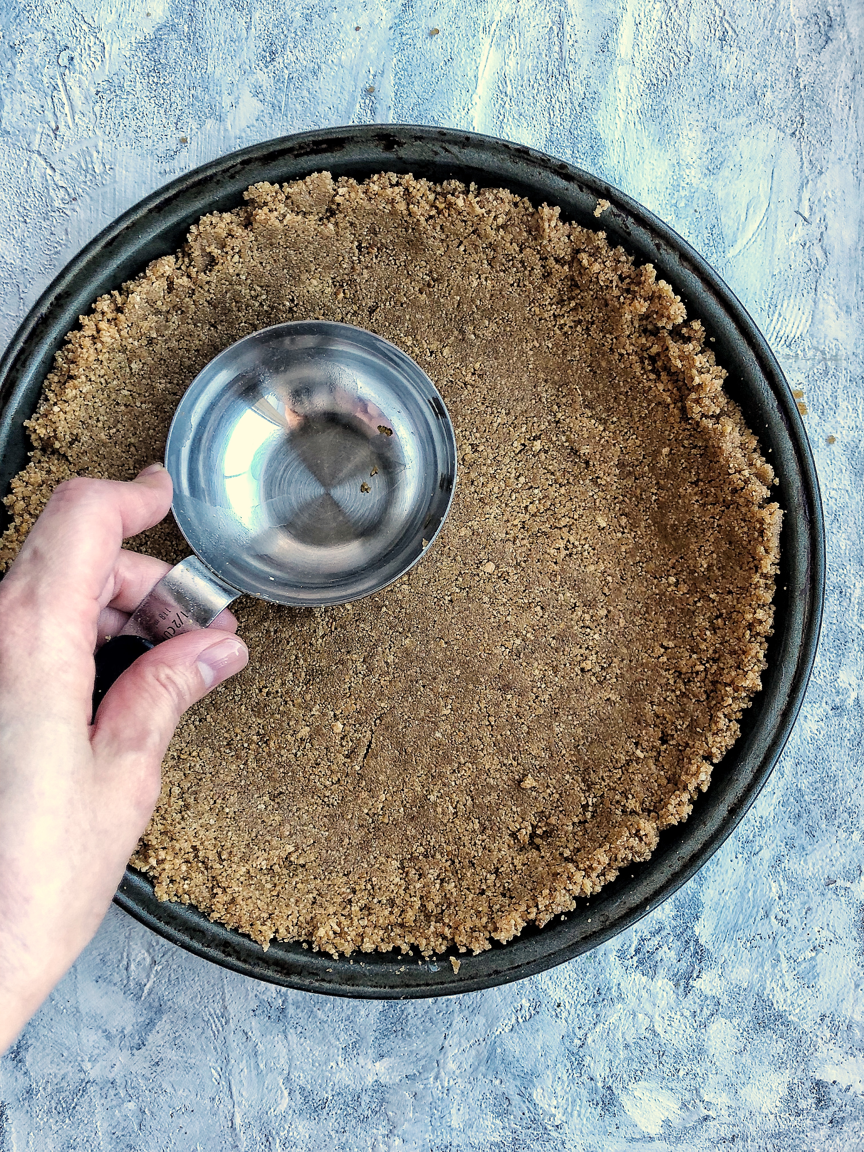 graham cracker crust in pie tin with hand holding measuring cup