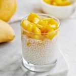 glass cup of coconut sago with mango topping on marble cutting board and mangoes in background