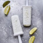 two coconut lime popsicles on gray sheet pan with limes