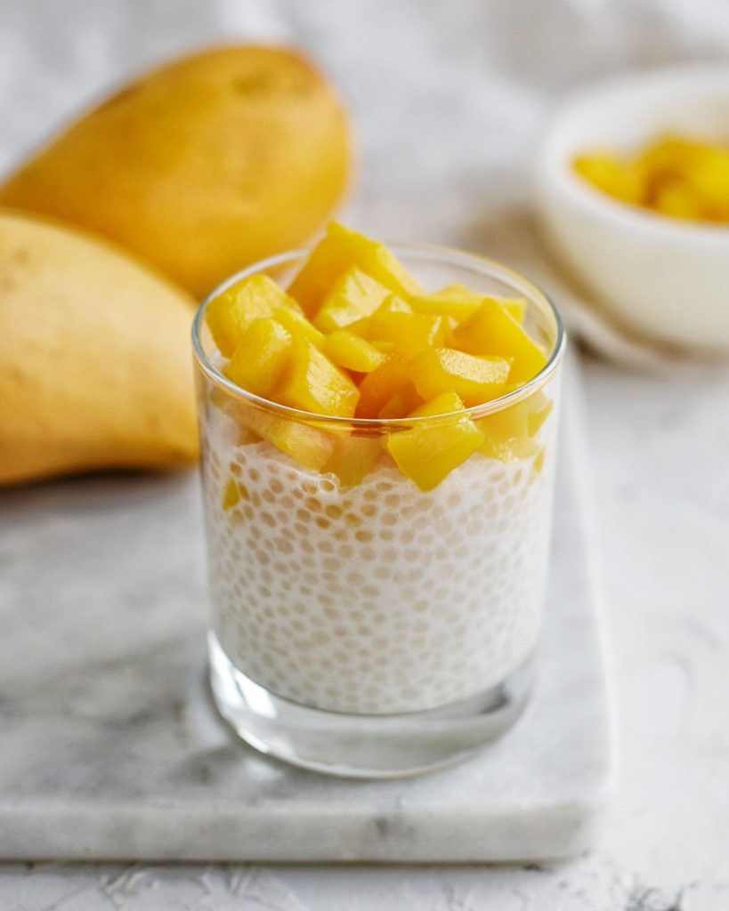 glass cup filled with coconut sago and mango topping with mangoes in background