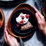 two hands on plate of chocolate almond cake with whipped cream and raspberries