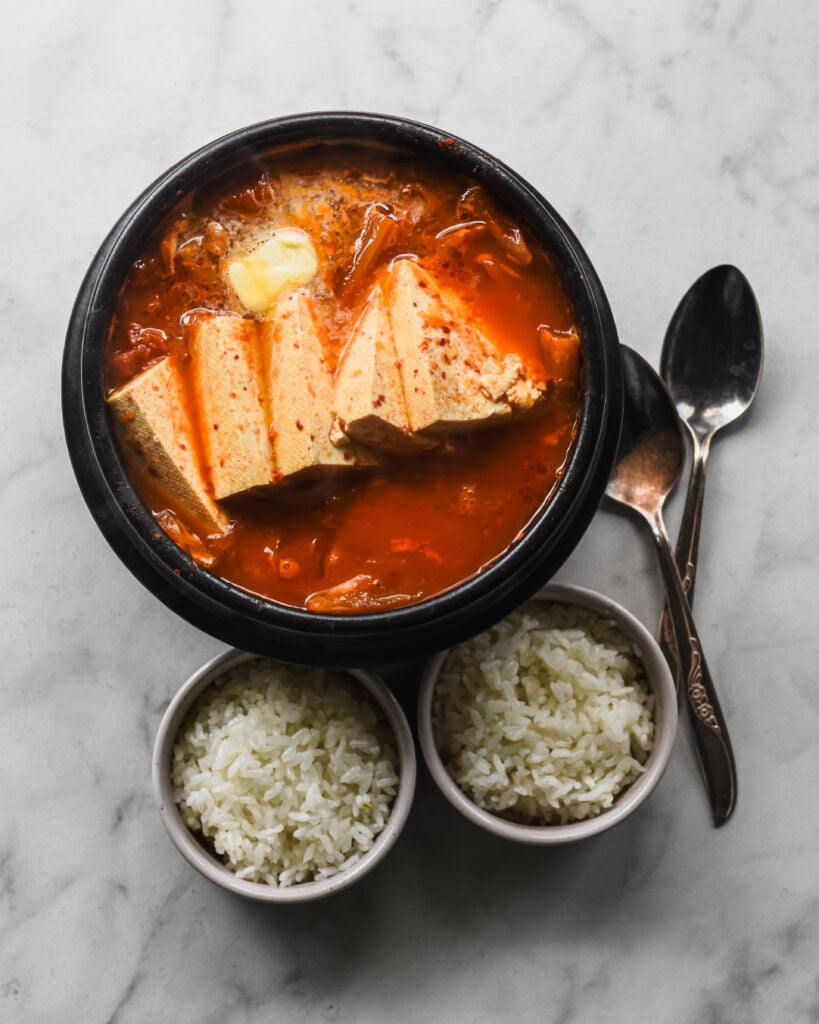 kimchi jjigae in korean clay pot with two bowls of rice and two spoons